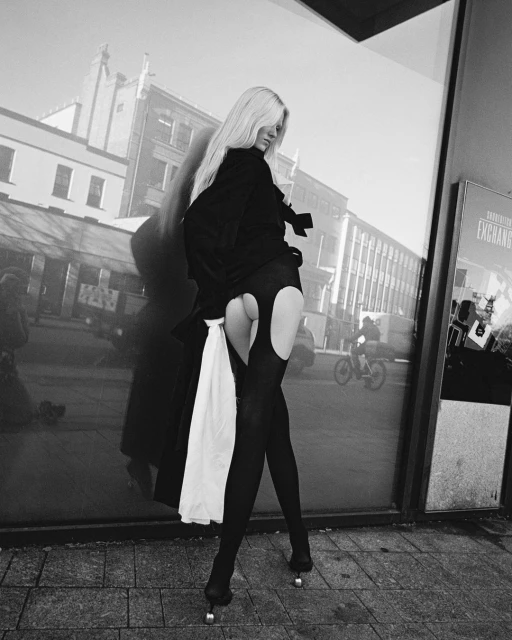 A person with long blonde hair poses in a city setting, wearing dark clothing and high heels, with a city scene reflected in glass.