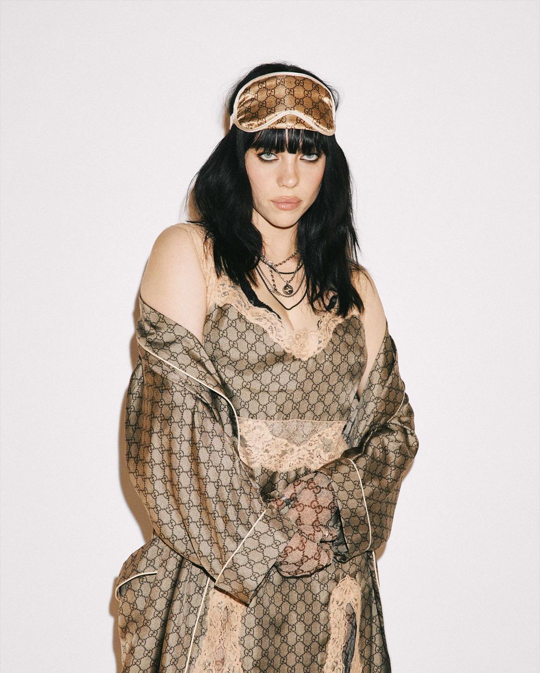 Portrait of a woman wearing a Gucci robe and sleep mask with a neutral expression against a white backdrop in a studio setting.