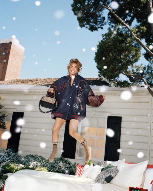 Adult woman in a letterman jacket jumps on a white bed outside a clapboard house with gifts during snowfall in a lifestyle shot.