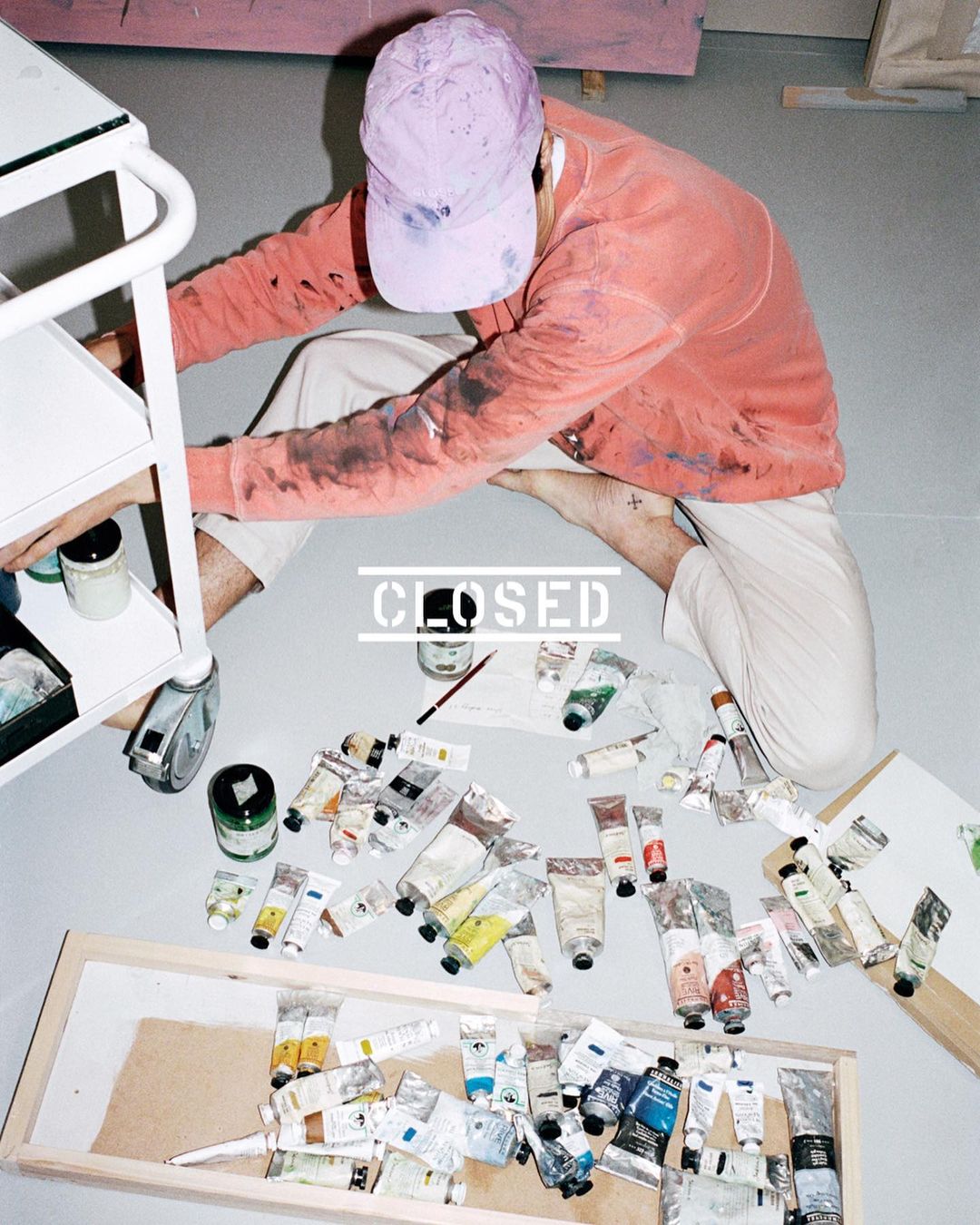 Overhead shot of an artist on the floor surrounded by paint tubes, wearing a cap and coral sweatshirt, with a supply cart nearby.