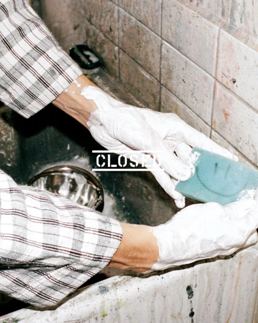 A person wearing gloves and a plaid shirt cleans a stainless-steel sink with a blue sponge, showcasing a focus on domestic tasks and cleanliness.