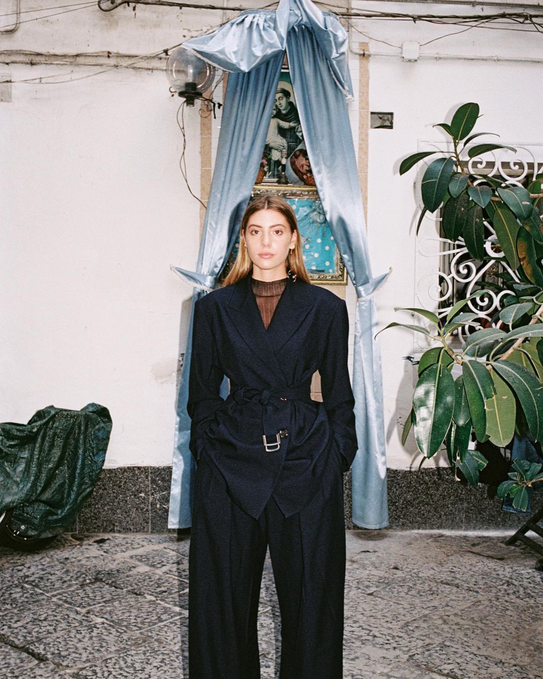 A woman in a dark suit poses in an outdoor setting with a shrine in the background, creating a high-fashion editorial look.
