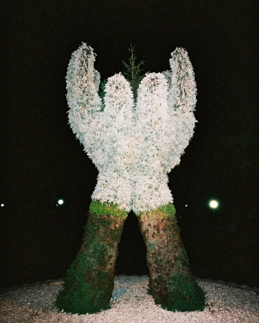 Topiary sculpture of hands supporting a small evergreen tree at night, set against a dark sky with distant lights.