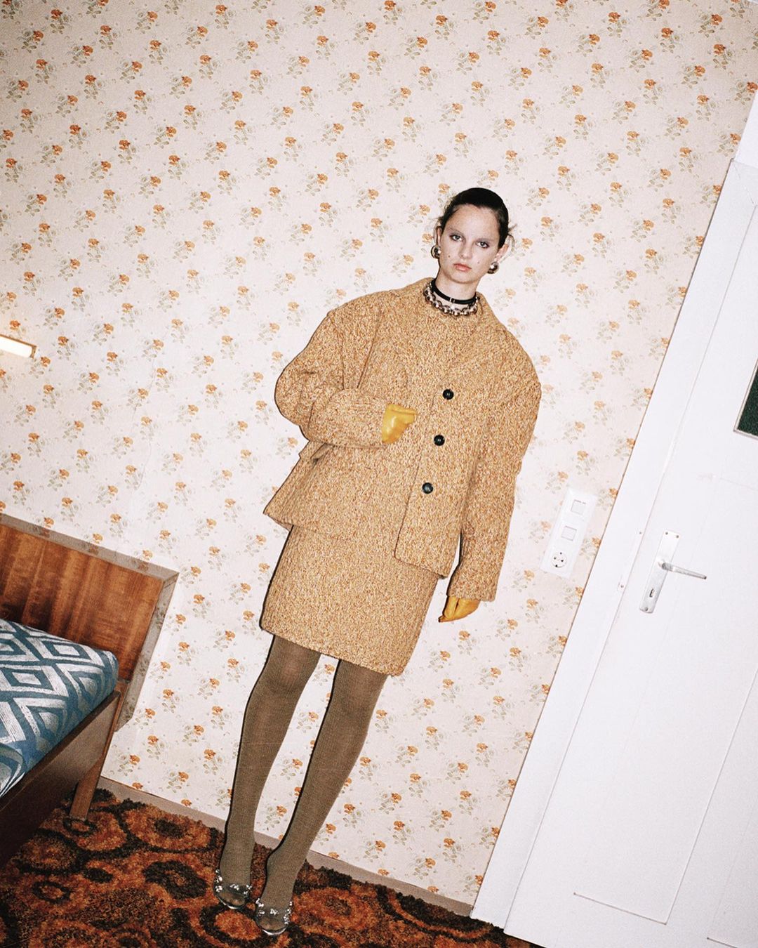 Woman in a tan skirt suit and sheer tights poses in front of a floral wallpaper and partially visible bed in a quirky room.