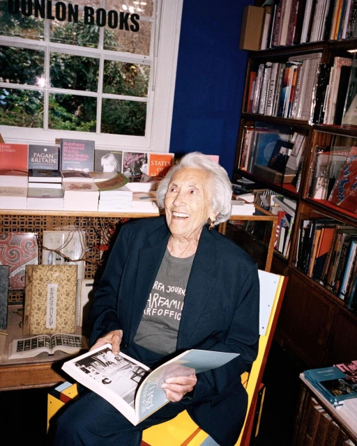 Elderly woman with white hair smiles while reading a book in the Dunlon Books bookstore, seated near bookshelves and a window.