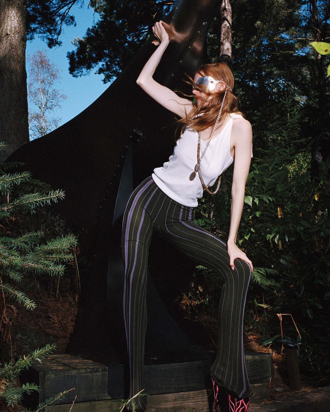 Fashionable redhead model poses in pinstripe pants and a white tank top, accessorized with jewelry, in an outdoor wooded setting.