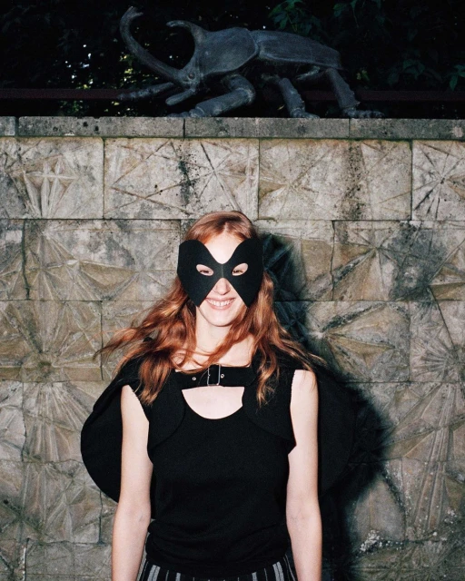 Young woman wearing black mask and top, smiling, stands against tiled wall with a large insect sculpture in the upper background.