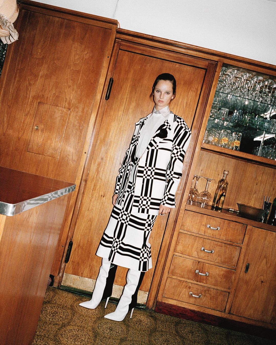 Woman in a black and white checker patterned coat and white boots stands in an indoor setting near wooden cabinets and countertops.