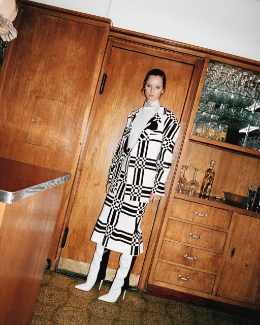 Woman in a black and white checker patterned coat and white boots stands in an indoor setting near wooden cabinets and countertops.
