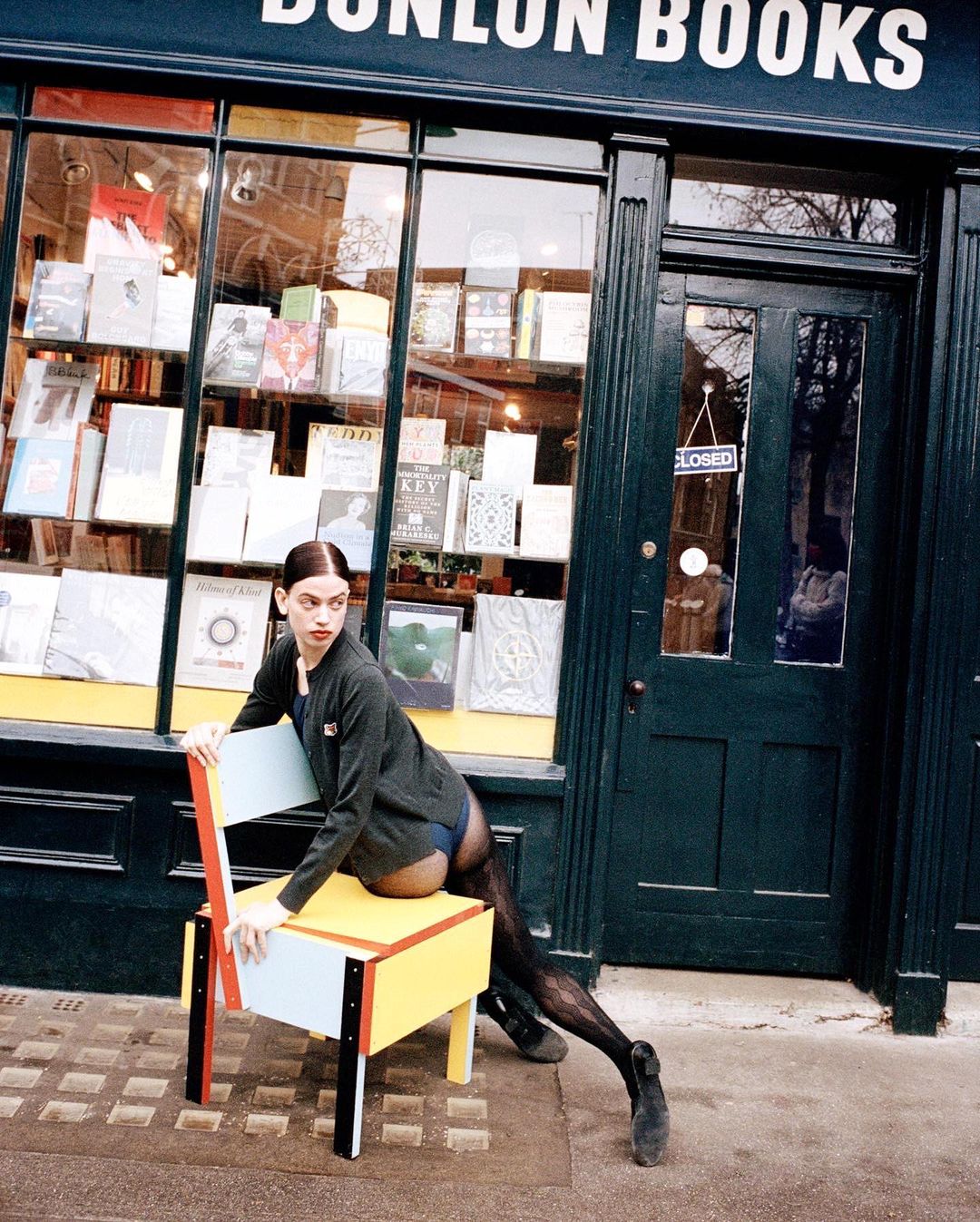 Woman in grey cardigan poses on a yellow chair outside of Dunlun Books, books visible through window, medium shot, natural light.