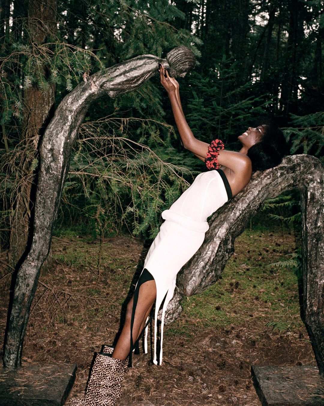 A fashion shot featuring a Black woman in a white dress, posing with a tree in a forest. She's styled with a red neck piece and animal print boots.