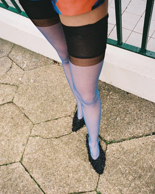 View of a woman's legs wearing sheer blue stockings and black high heels, standing on a textured gray concrete surface outdoors.