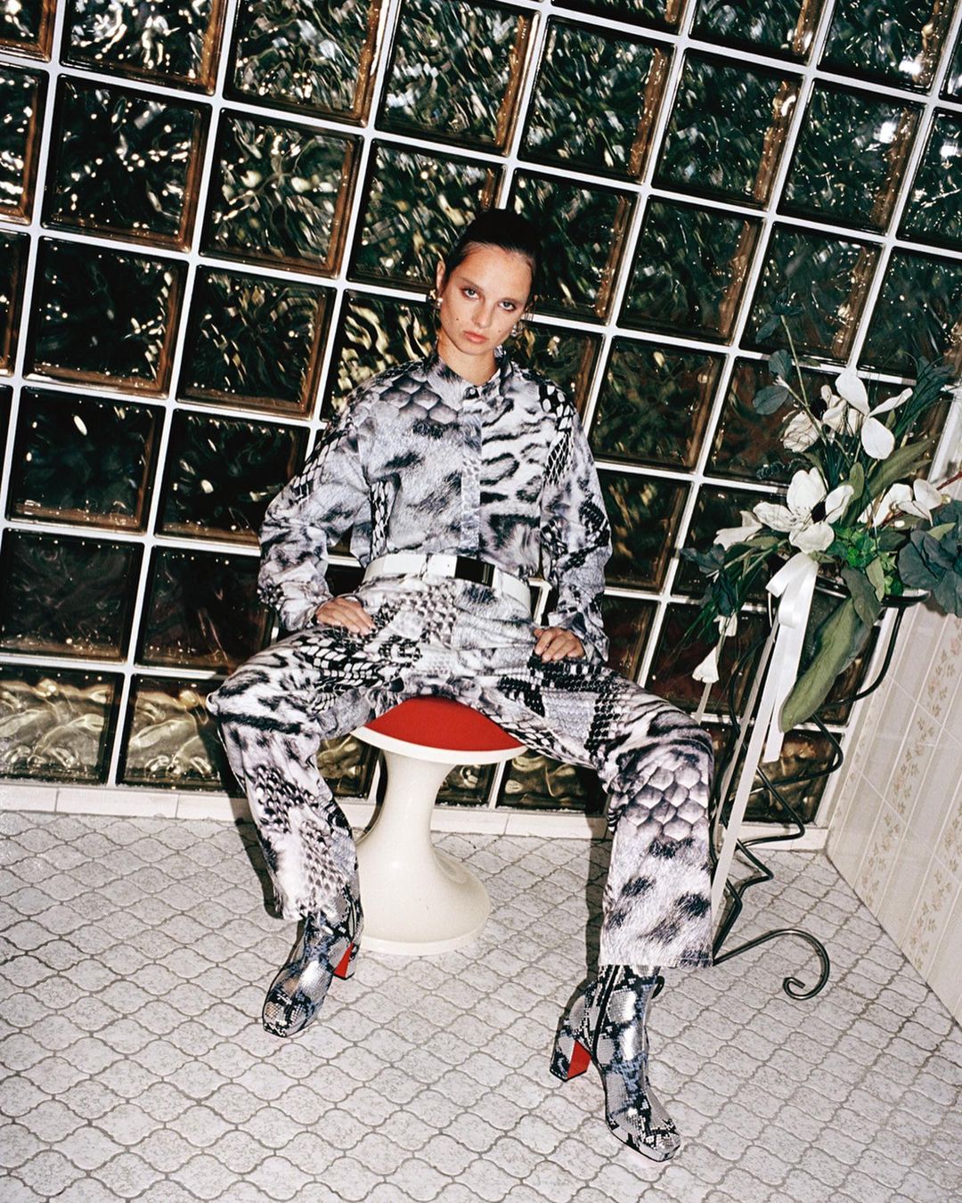 A woman in patterned clothing sits on a stool, facing the camera with a serious expression, in a room with a tiled floor and glass block wall.