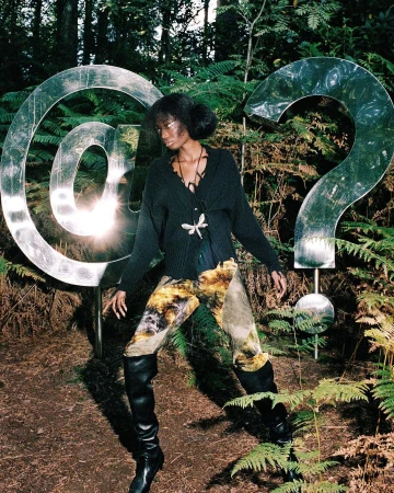 A woman poses between metal @ and ? symbols in a forest setting, wearing a cardigan, tie-dye pants, and high-heeled boots.