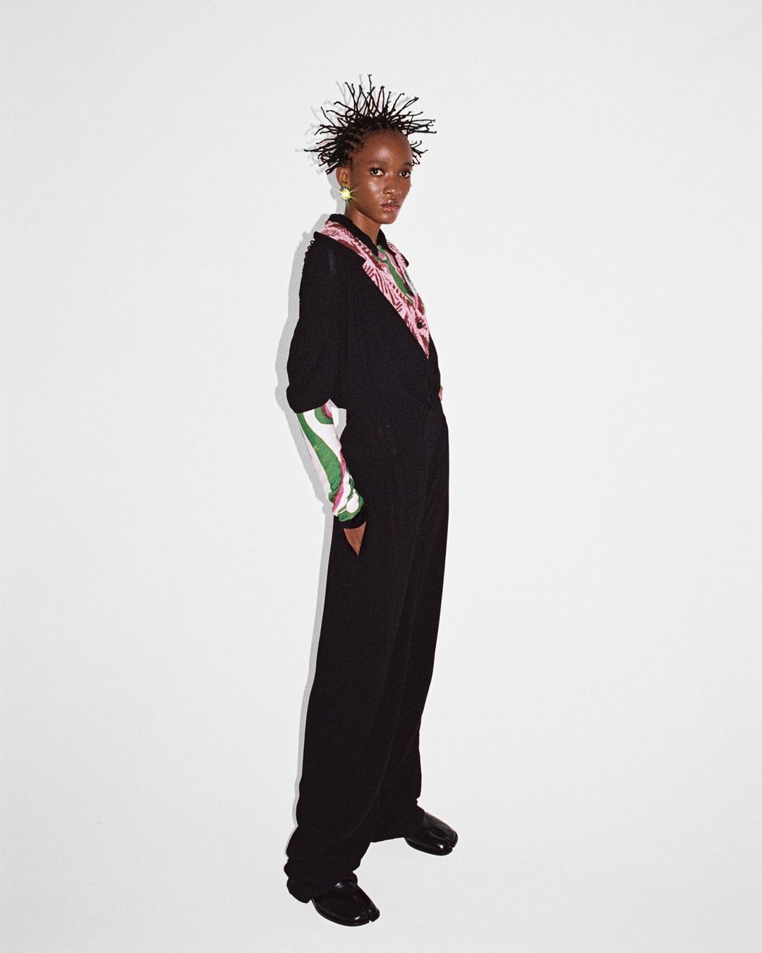 A slim Black model with short spiked hair poses in a black jumpsuit with green and pink accents against a white backdrop in studio lighting.
