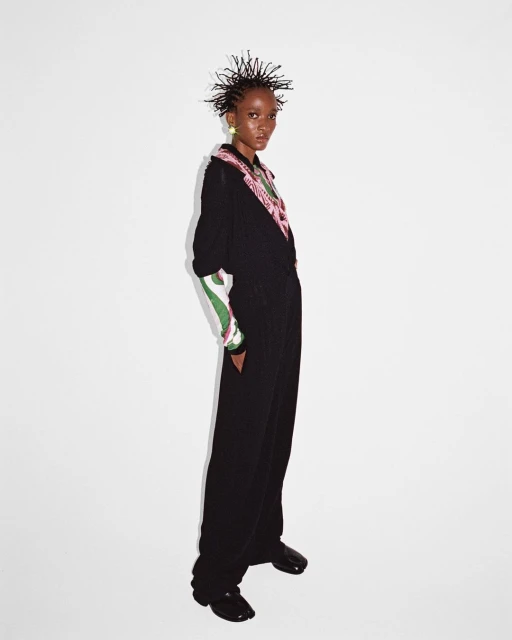 A slim Black model with short spiked hair poses in a black jumpsuit with green and pink accents against a white backdrop in studio lighting.