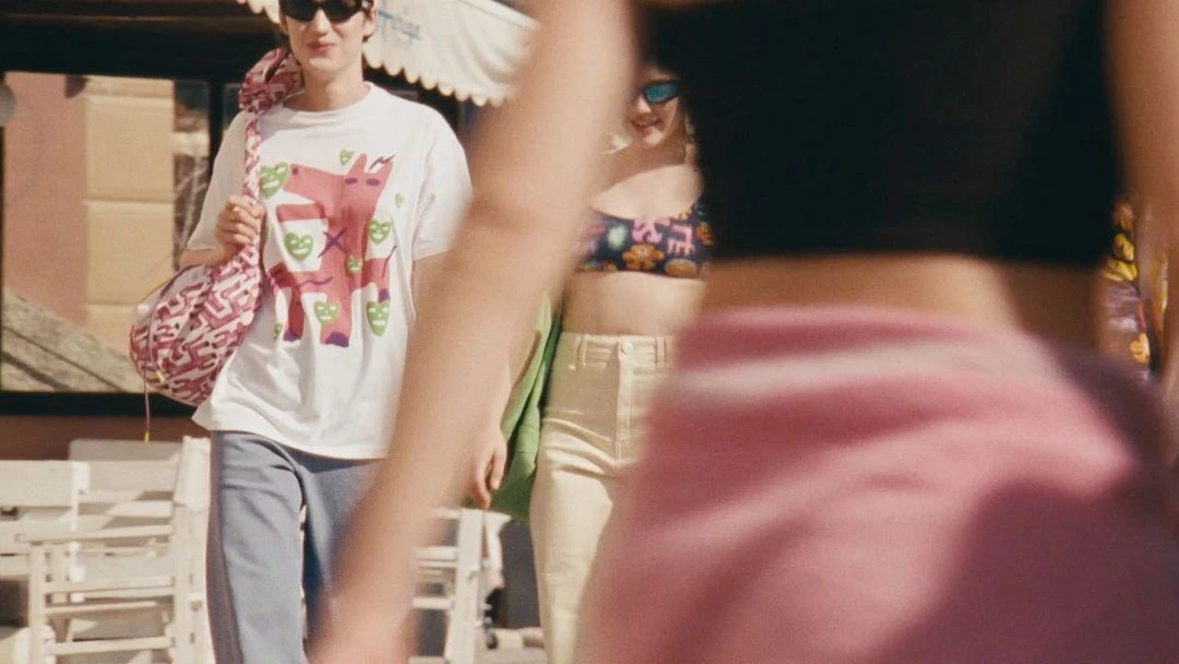 Three people walk outside on a sunny day; one wears a graphic tee and jeans, another a bikini top and jeans, and one is out of focus.