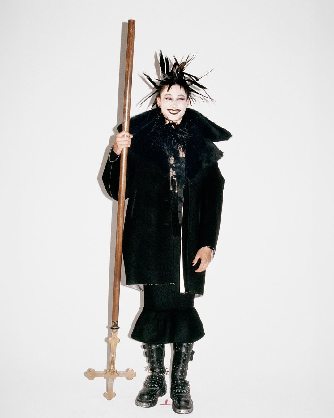 Person in black coat and spiked headdress holds wooden cross on white background. Fashion forward, editorial, stylized shot.