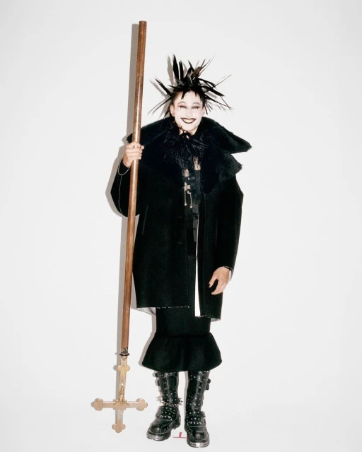 Person in black coat and spiked headdress holds wooden cross on white background. Fashion forward, editorial, stylized shot.