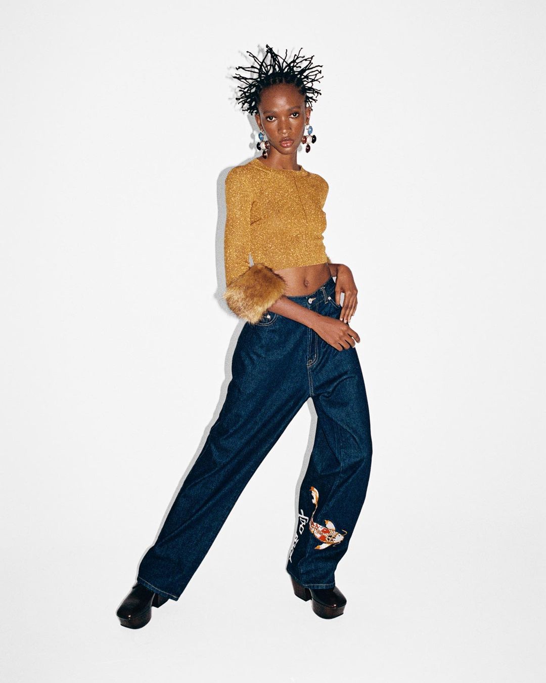 Fashionable young Black woman poses in a gold crop top with fur cuffs and wide-legged jeans with koi fish embroidery on a white backdrop.