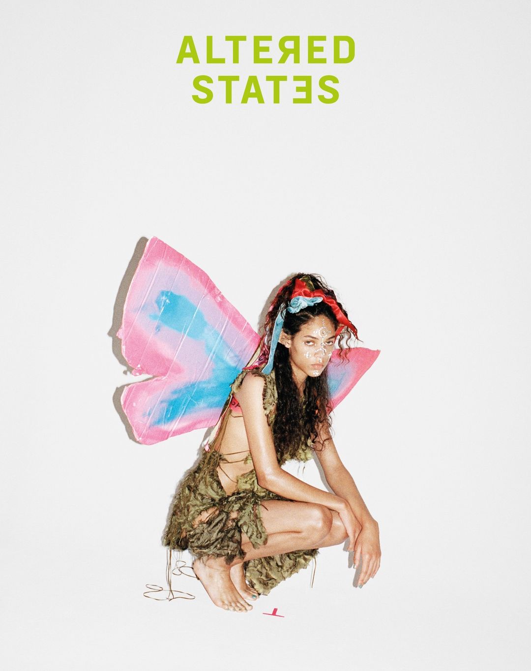 Young woman with dark hair poses in fairy costume with pink and blue wings against a white background under bright lighting.