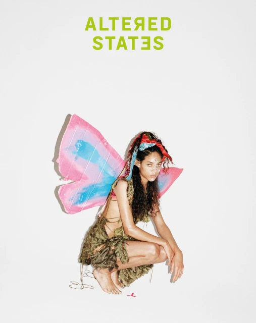 Young woman with dark hair poses in fairy costume with pink and blue wings against a white background under bright lighting.