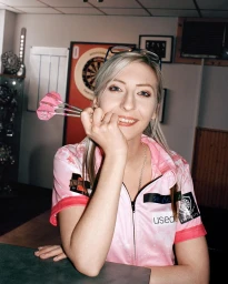 Blonde woman in a pink sports shirt holding three darts near her face, photographed by Anton Gottlob