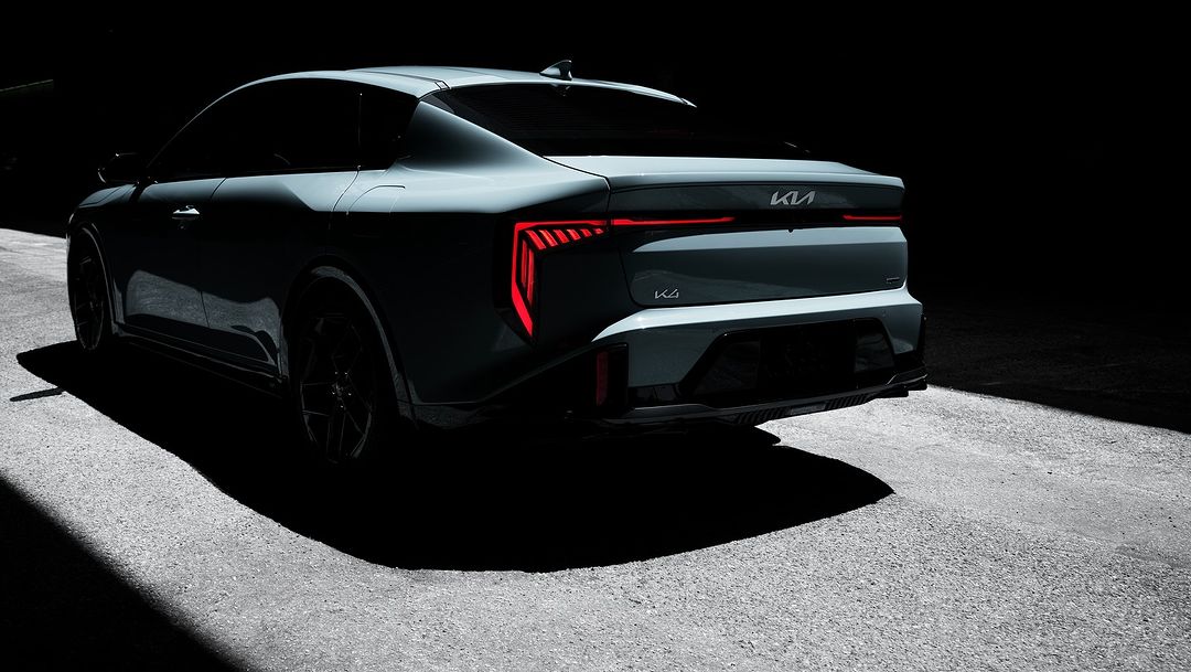 Rear view of a gray Kia K4 car parked on asphalt with tail lights illuminated, set against a dark background.