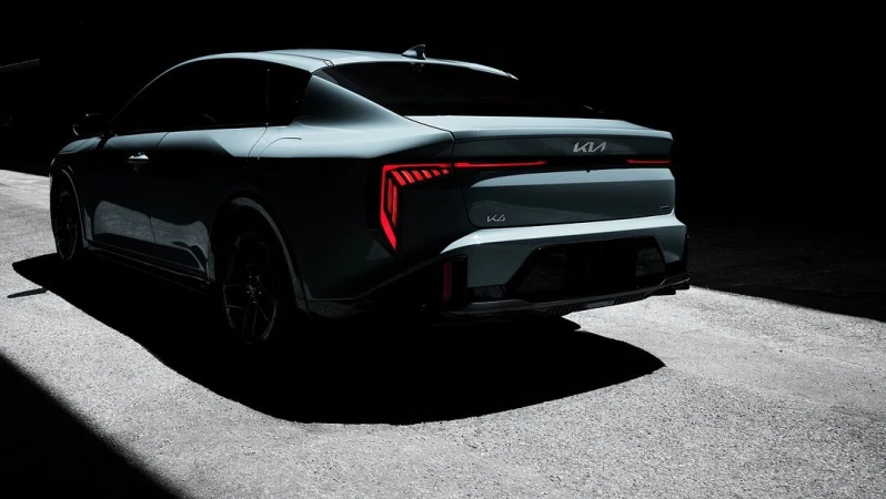 Rear view of a gray Kia K4 car parked on asphalt with tail lights illuminated, set against a dark background.