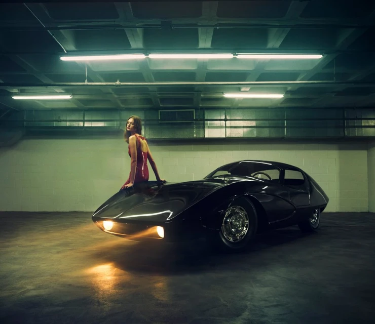 Adult woman in a red dress sits on the hood of a black classic car in an indoor parking garage with fluorescent lighting.