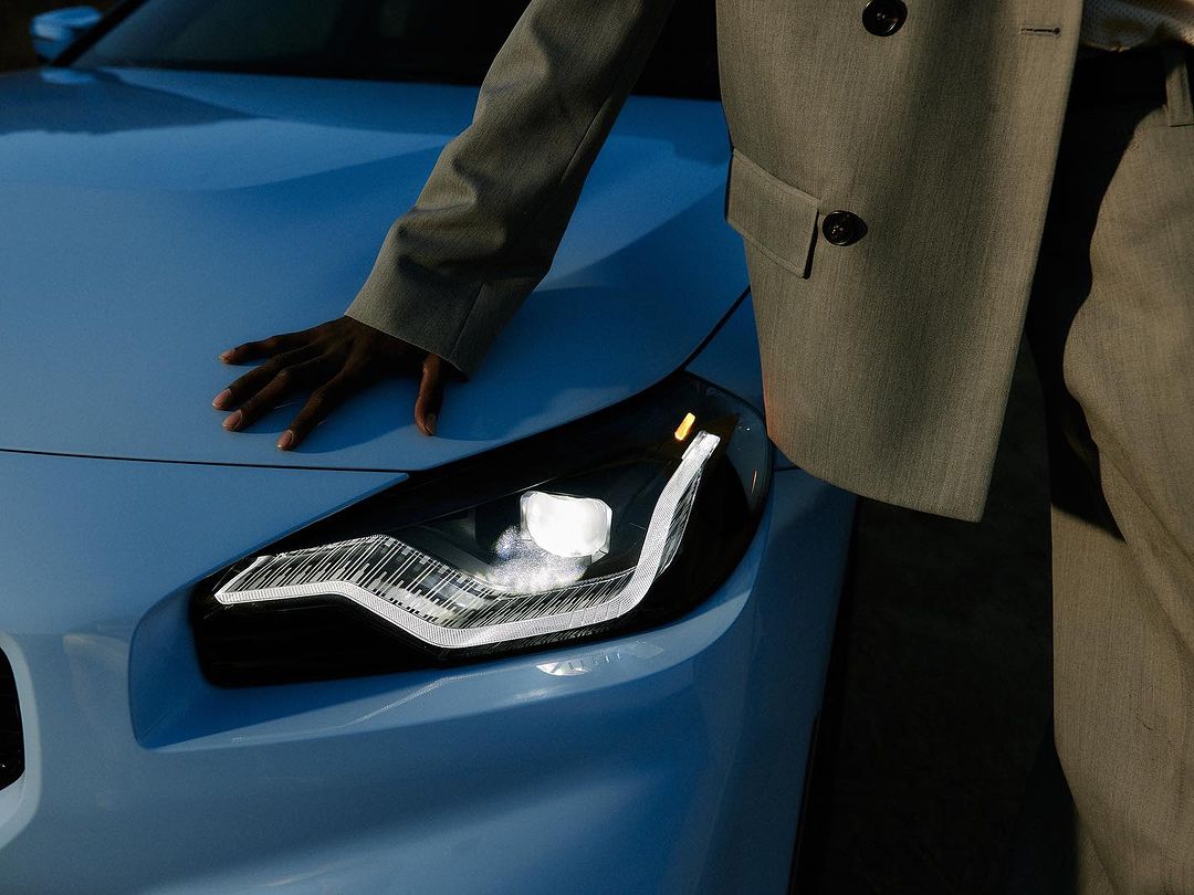 Close-up of a light blue car with its modern headlight illuminated, an African-American person wearing a suit rests their hand on the hood.