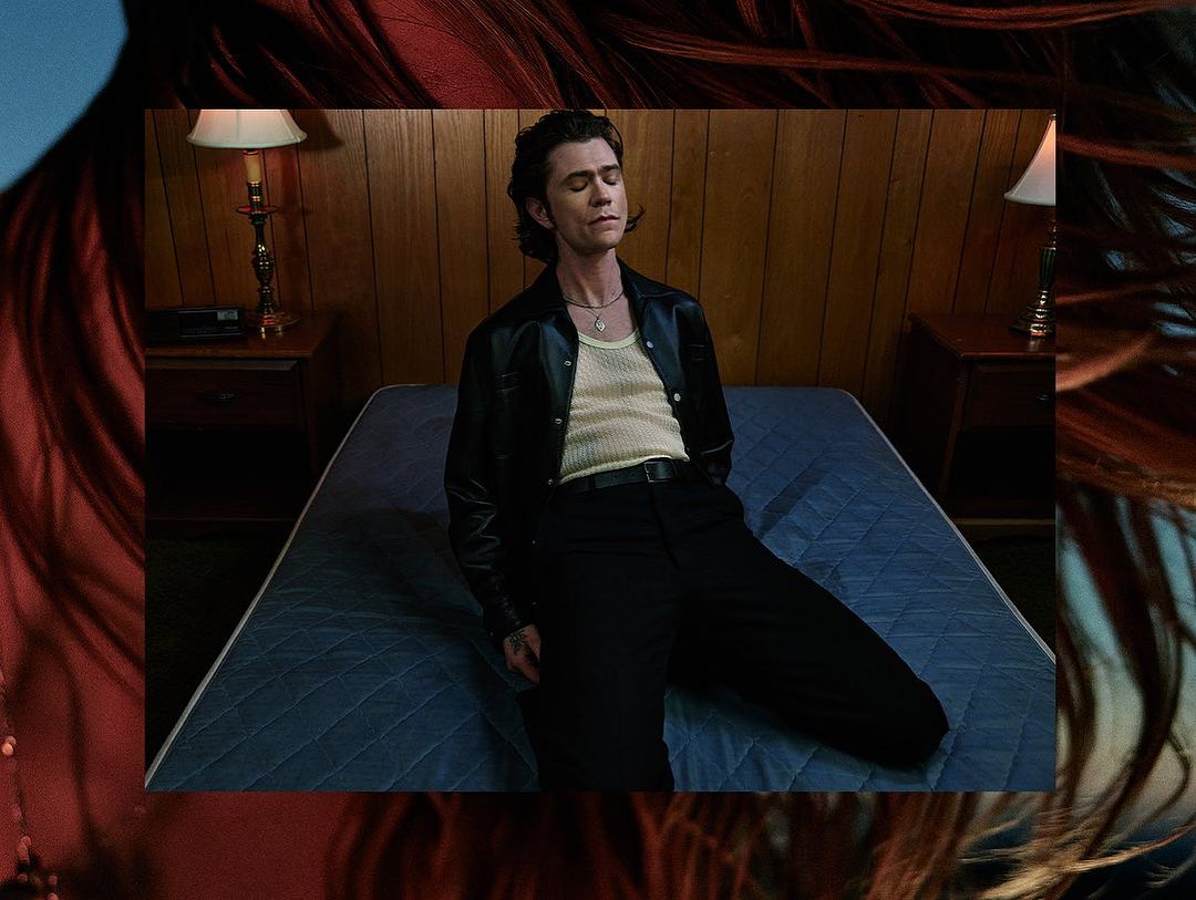Portrait of a male model in black leather jacket sitting on a bed with wood paneled walls in the background, stylized portrait.