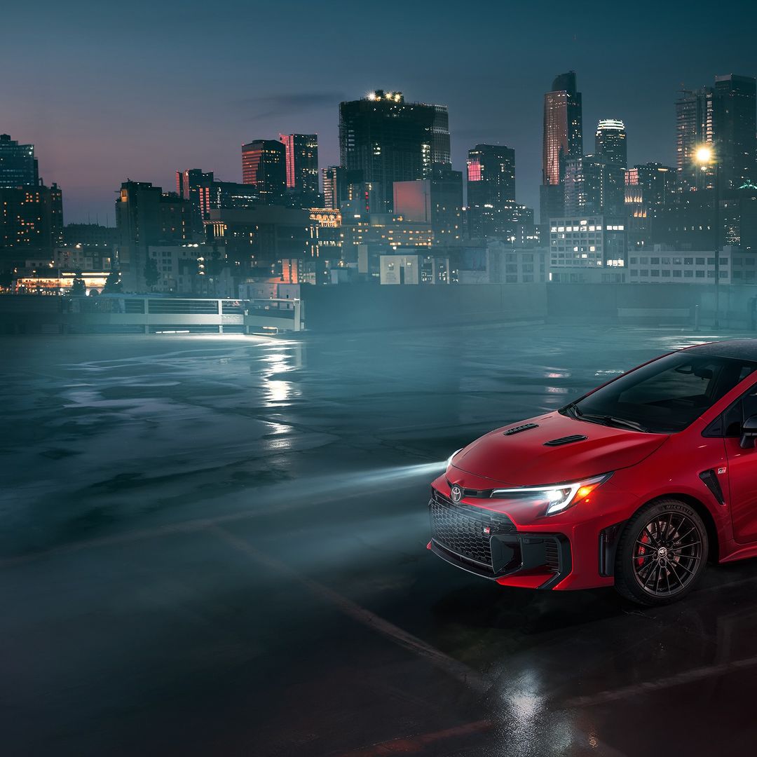 Red Toyota GR Corolla sports car parked on a wet surface at night with city skyline backdrop, headlights on, creating a moody atmosphere.