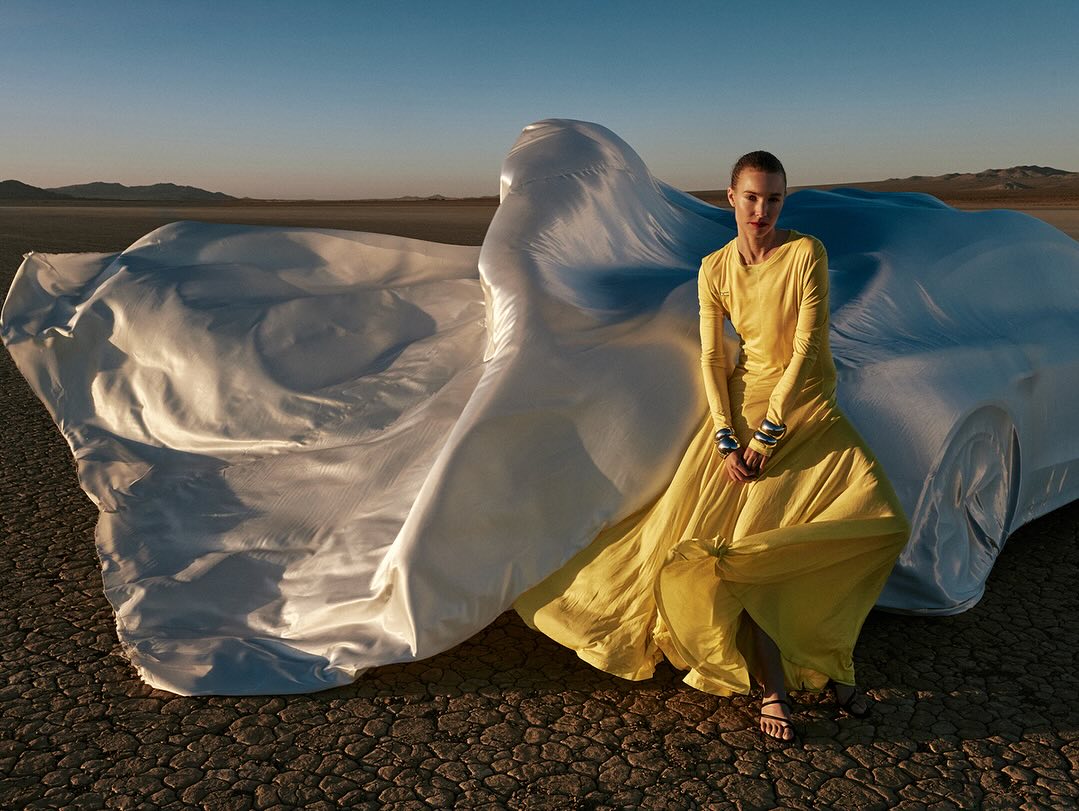 Fashion shot of a woman in a yellow dress posing in a desert environment with a covered car, artistic composition, and neutral color palette.