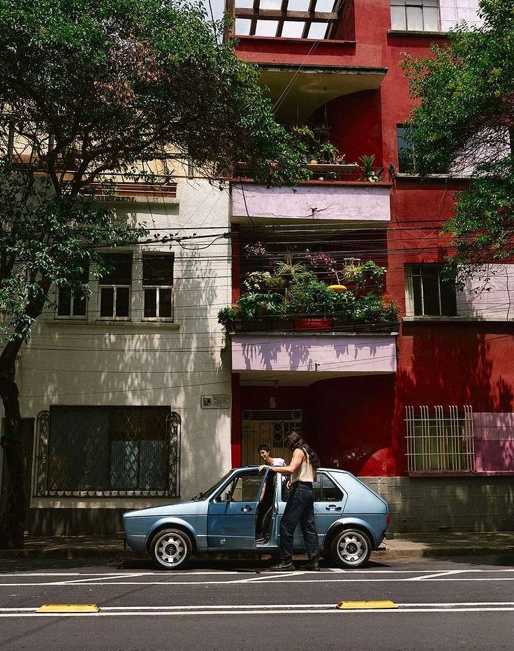 Adult female exiting a light blue car on a city street next to a white building with plants and flowers on a balcony, sunny day.