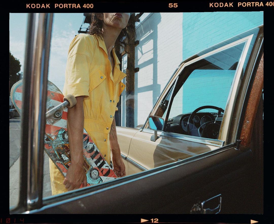 A person in a yellow jumpsuit stands next to a vintage car, holding a skateboard against a light blue building and a sunny sky.