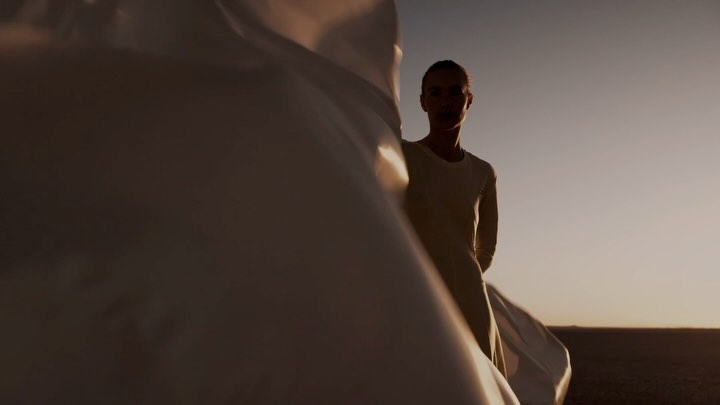 Woman in flowing dress stands against a desert background under a clear sky, bathed in warm, golden light during golden hour.