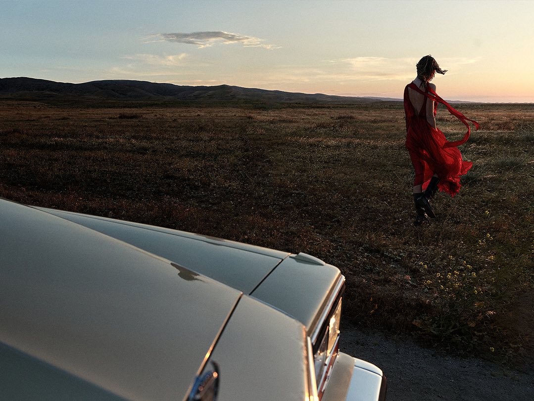 Adult woman in a red dress walks across a grassy field at sunset near a vintage car, with rolling hills and a sky in the background.