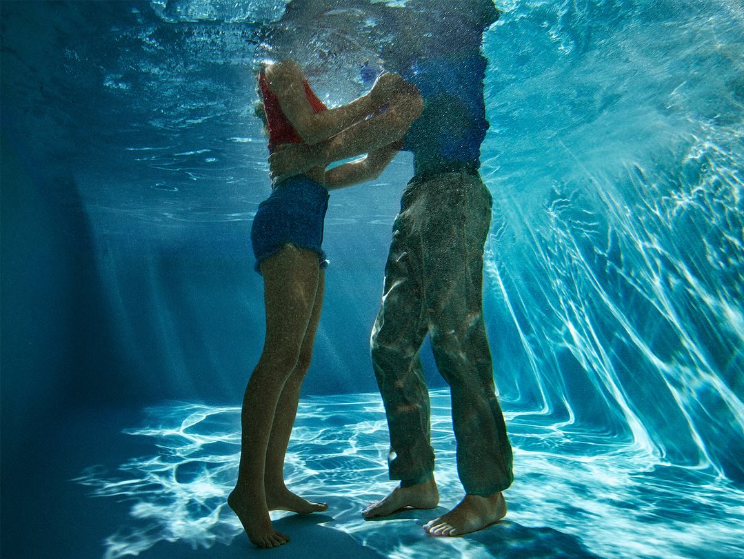 Underwater view of a couple embracing in clear blue water. The woman wears a red top and blue shorts, while the man is in a blue shirt and pants.