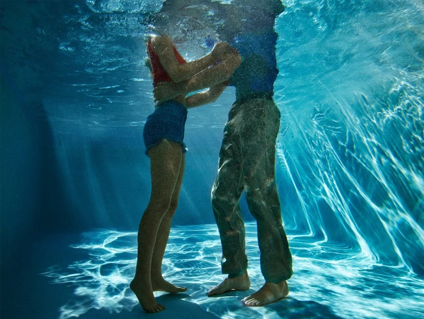 Underwater view of a couple embracing in clear blue water. The woman wears a red top and blue shorts, while the man is in a blue shirt and pants.