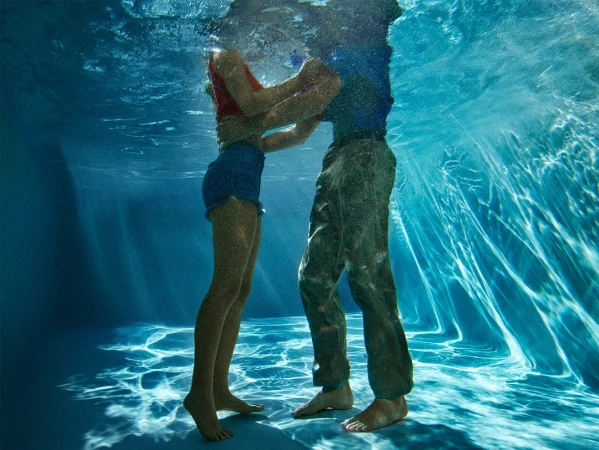 Underwater view of a couple embracing in clear blue water. The woman wears a red top and blue shorts, while the man is in a blue shirt and pants.