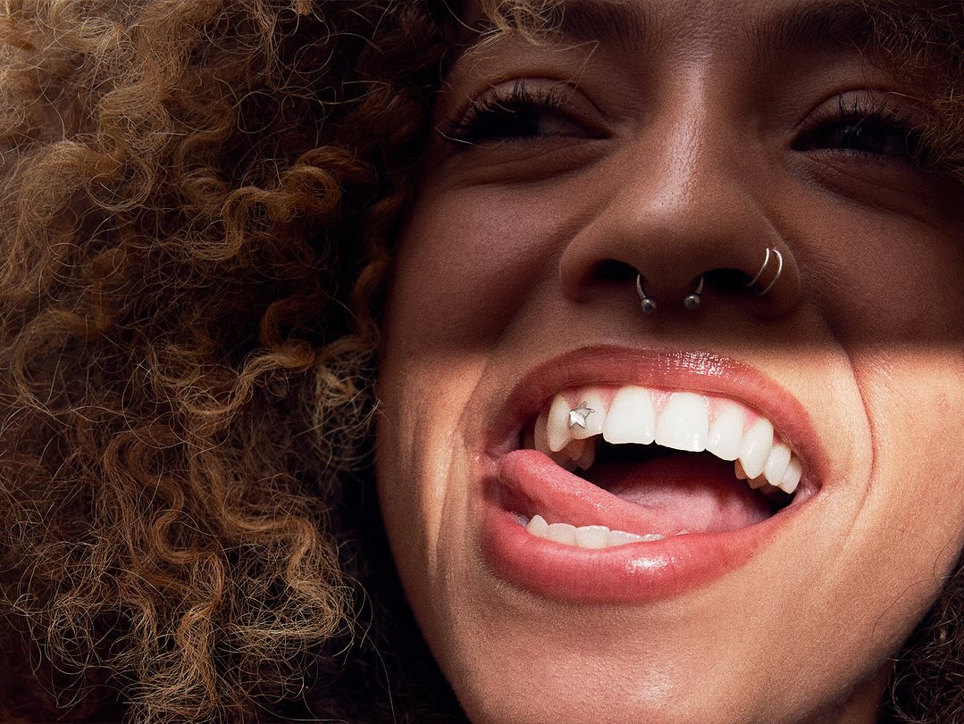 Close-up of a young woman smiling with a star tooth gem, curly hair, and facial piercings with bright, natural lighting.