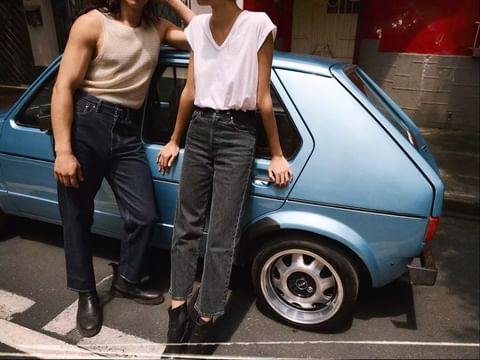 A couple poses near a light blue compact car on a city street. The woman wears a white shirt and black jeans, while the man wears a tank top.