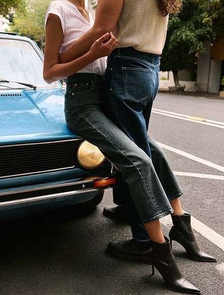 Two people standing close by a light blue vintage car on a city street, embracing on an overcast day in casual outfits.