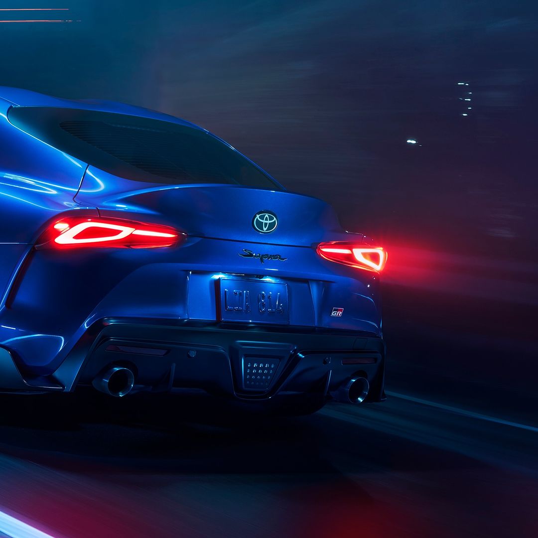 A sleek blue Toyota Supra sports car is captured from the rear, showcasing its modern design and illuminated tail lights at twilight.
