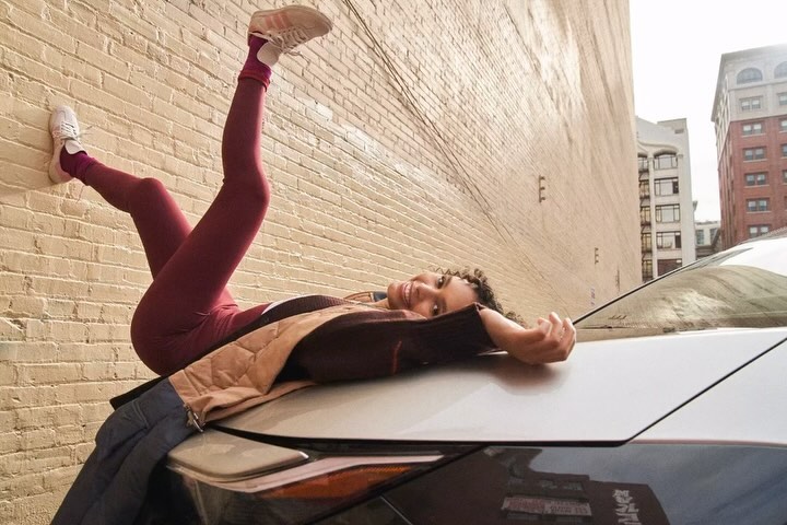 A smiling woman with curly hair poses on the hood of a car with her legs up on a brick wall in an urban environment.