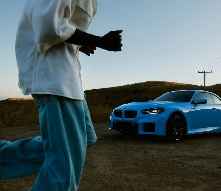 Man in white shirt and loose pants running in a field, a light blue BMW M2 coupe is parked nearby against a blue sky backdrop.