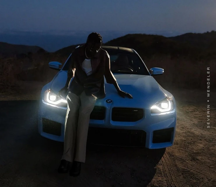 An adult male leans on the hood of a light blue BMW car with illuminated headlights at dusk with mountains in the distance.