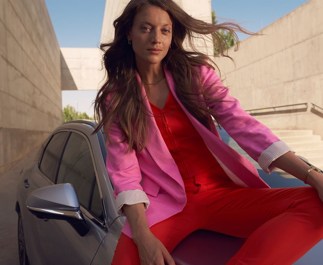 Brunette woman in a pink jacket and red pants sits on a gray car with a modern building in the background on a sunny day.