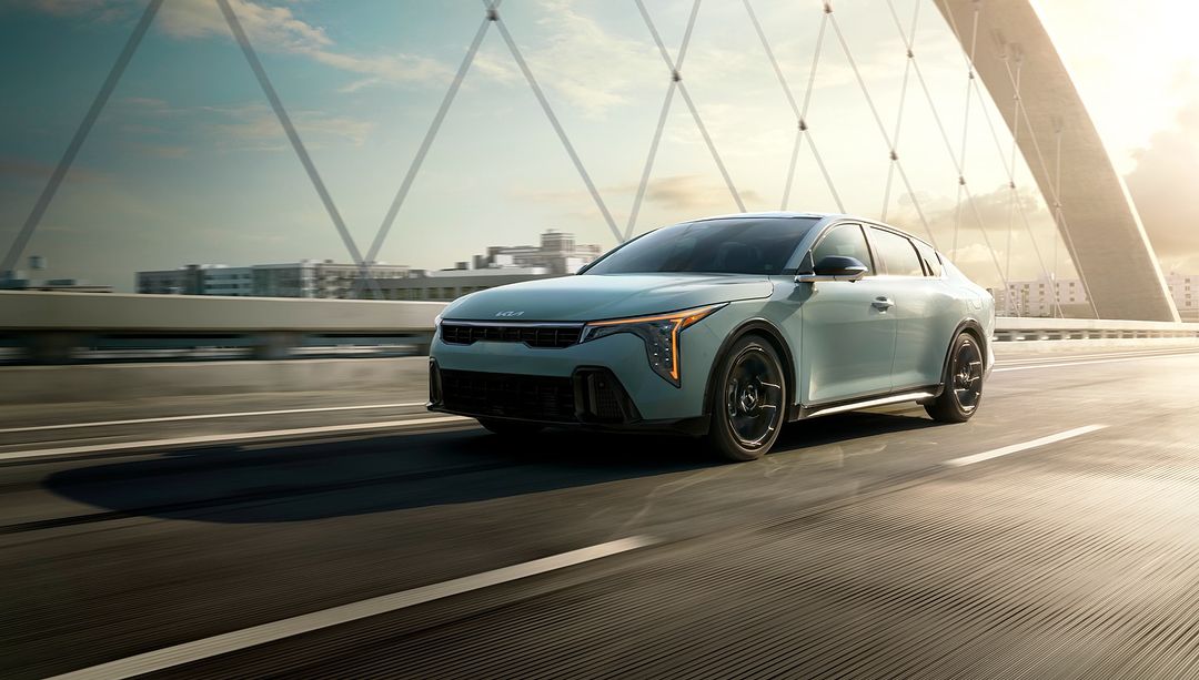 Modern light blue KIA EV5 electric car driving on a bridge, with a city skyline and bright sky visible in the background.
